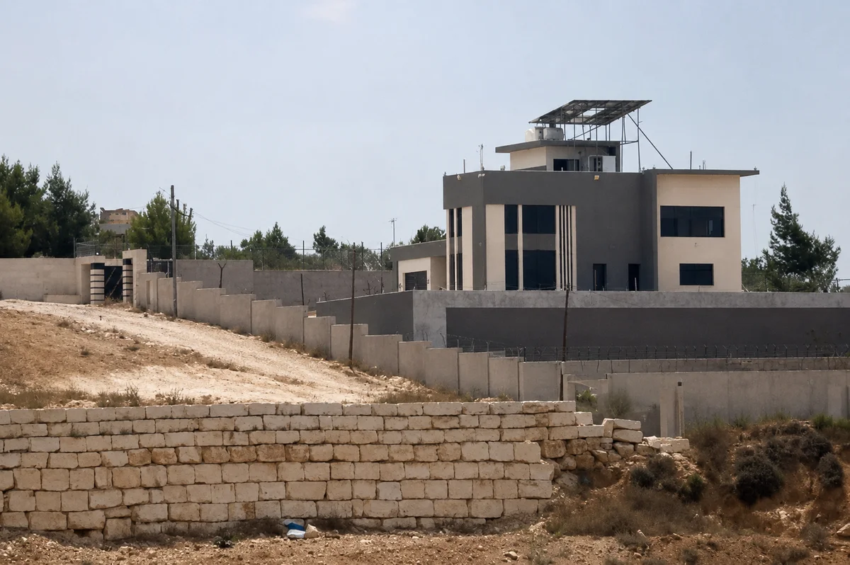 A realistic view of a large residential building and surrounding walls in the dry West Bank landscape.